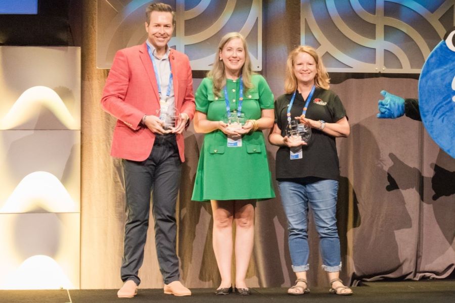 Three people stand on stage holding awards next to a blue higher education company mascot.