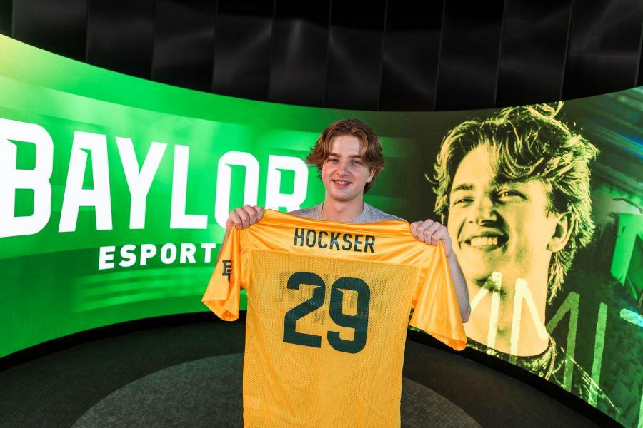 Colby "hockser" James hold a gold jersey with his name and number 29 with Baylor esports in the background.