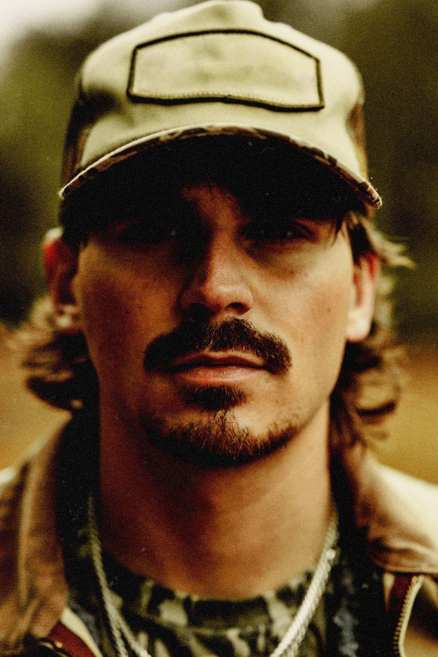 Country singer Tucker Wetmore wearing a billed cap, a camouflage shirt, a gold neckchain and collared jacket