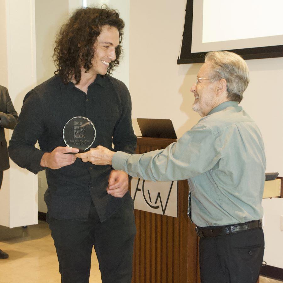 Playwright Jack Angelo Cummings dressed in black is handed circular crystal award for his new play from Preston Kirk.