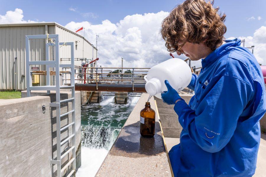 Man in a blue work jumpsuit pours water from a jug into a water testing bottle.