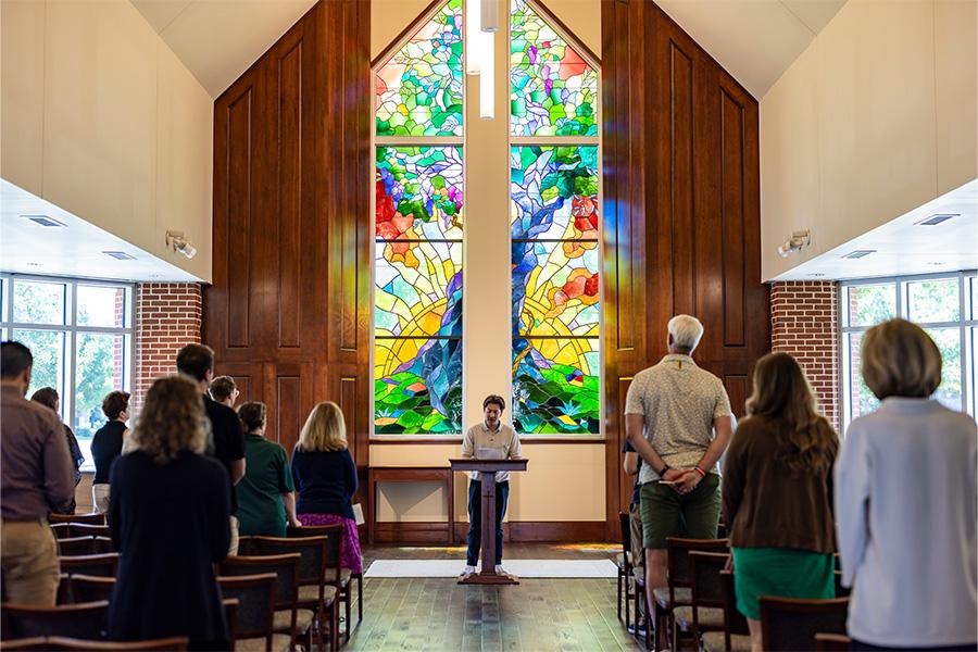 Minister speaks during a service in a chapel with colorful stained glass