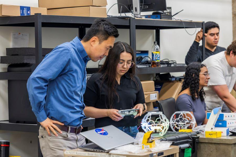 Professor in a long-sleeved blue shirt works with a student building a drone.