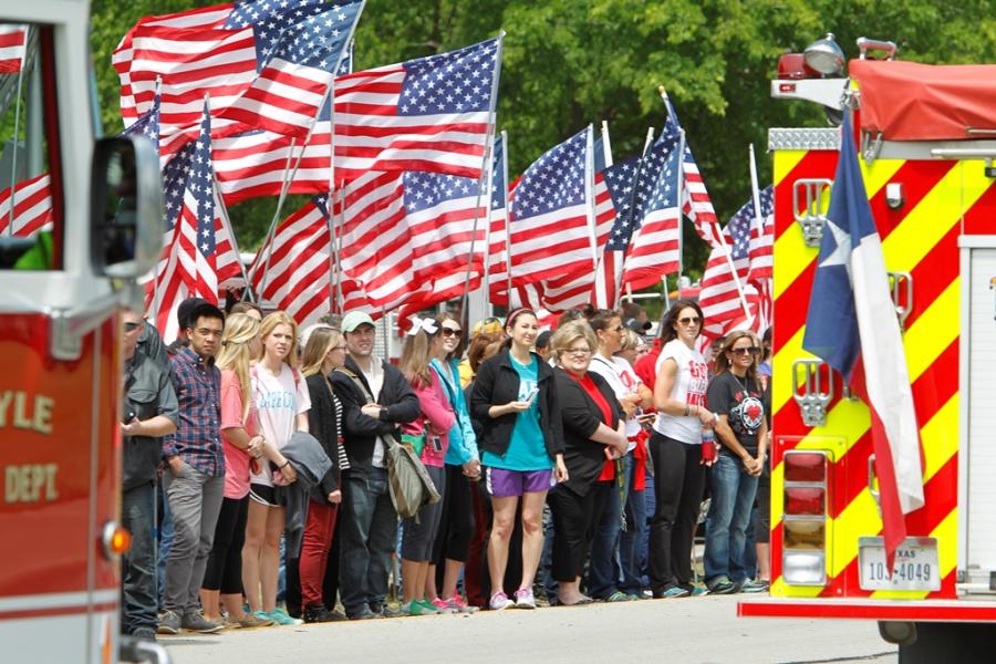 Photo of people holding American flags on the sides of streets as a parade procession passes.